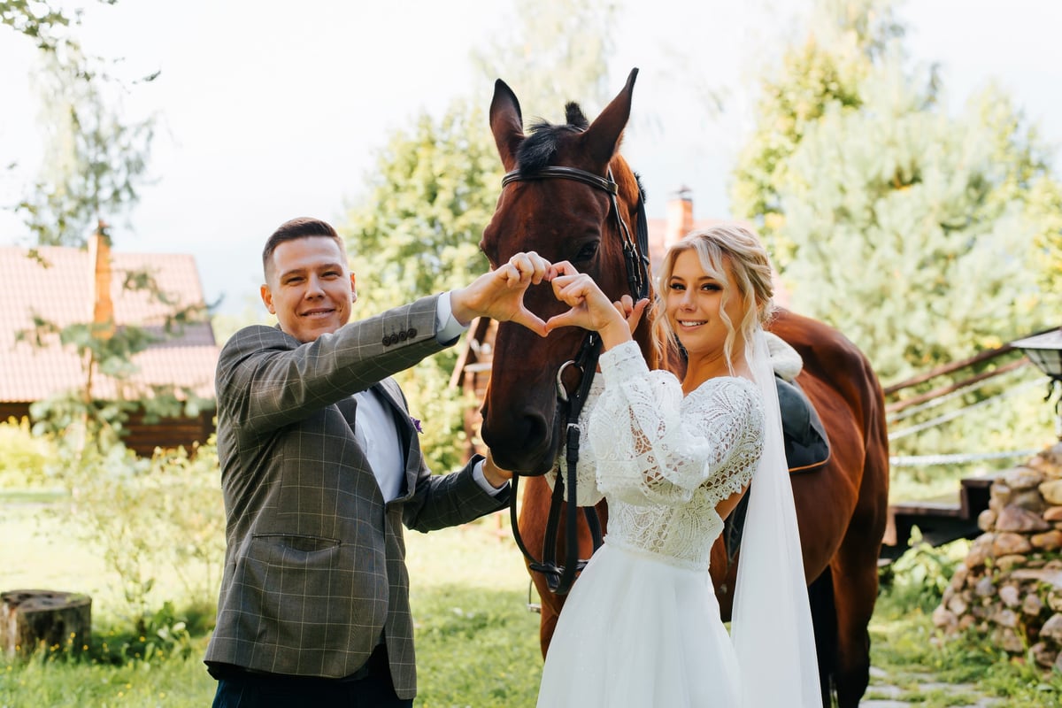 Portrait of happy young married couple with horse outdoors, smiling newlyweds folded their hands in shape of heart sign of love, bride and groom looking at the camera. Wedding day, marriage concept.