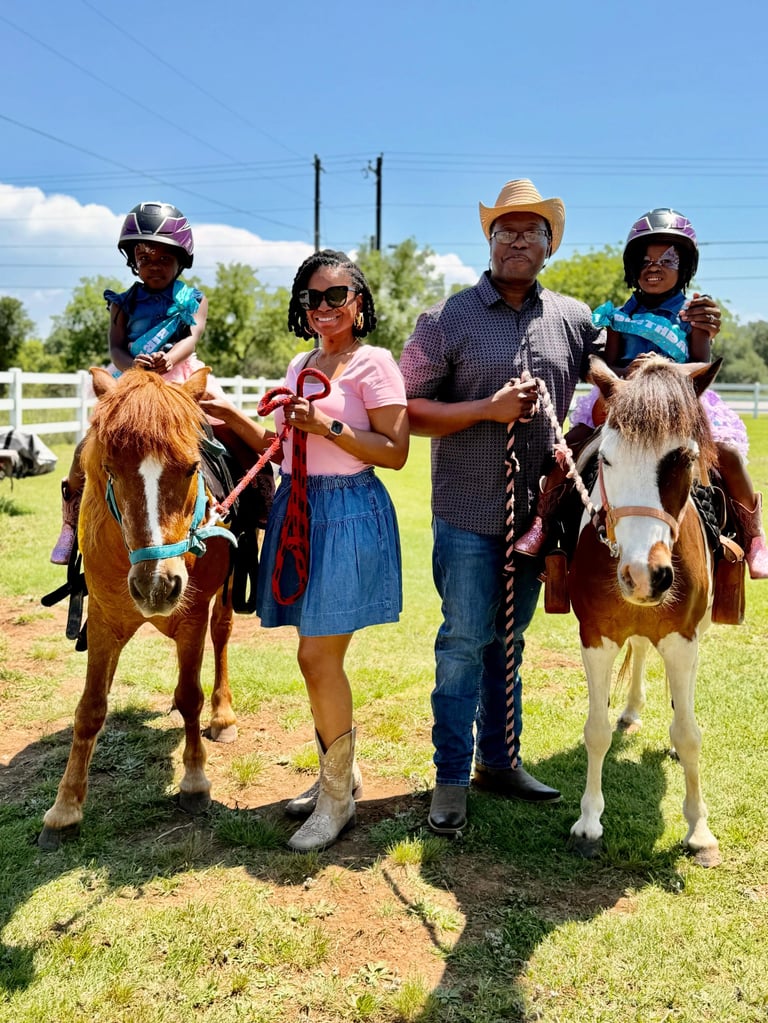 Four people wearing helmets standing with two small brown and white horses on a sunny day at a ranch with green grass and white fencing
